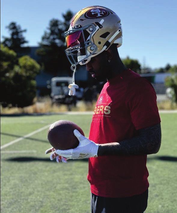 Love in a 49ers helmet and shirt holding a football on a practice field